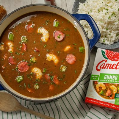 Stovetop Cajun Gumbo in a blue dutch oven featuring shrimp and sasuage next to a box of camellia brand gumbo roux base and a plate of white rice