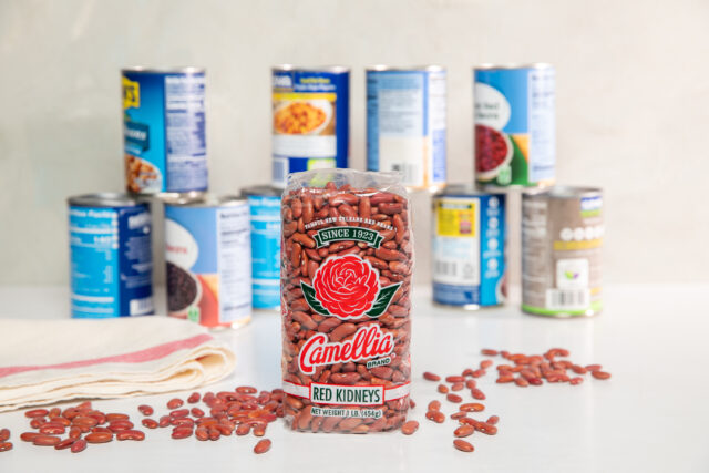 bag of Camellia Brand red kidney beans standing upright with 2 other brand's cans of red beans stacked in background, somewhat out of focus, with individual red beans loose and scattered around foreground