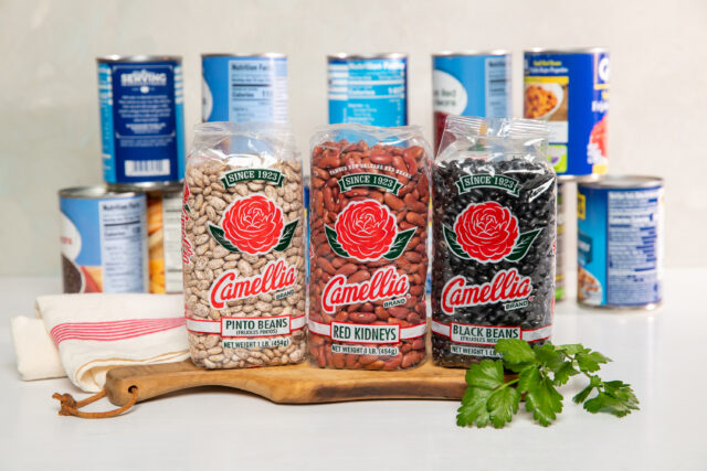 3 bags of dry Camellia Brand beans in front of row of various other brand's canned beans on white backdrop