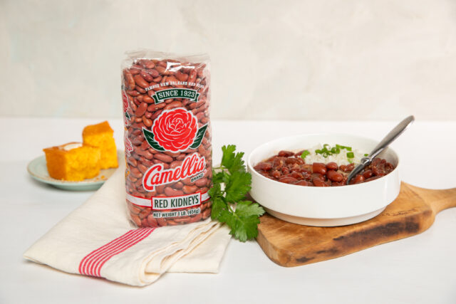 bag of Camellia Brand dry red beans upright next to wooden cutting board with a bowl of cooked red beans on top of it, with cornbread pieces in background