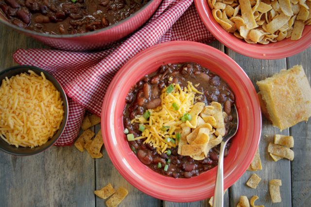 a bowl of 3 bean vegetarian chili next to frito chips, shredded cheese and a dutch oven of beans