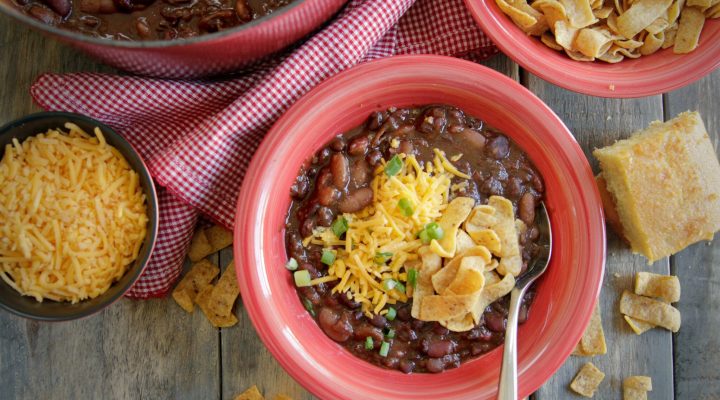 a bowl of 3 bean vegetarian chili next to frito chips, shredded cheese and a dutch oven of beans