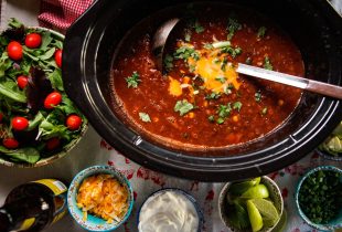 a slow cooker full of slow cooker lentil black bean taco soup next to a bunch of bowls with various toppings for the soup