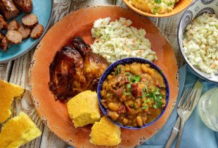 a plate of baked beans and bbq chicken with a side of coleslaw and corn bread