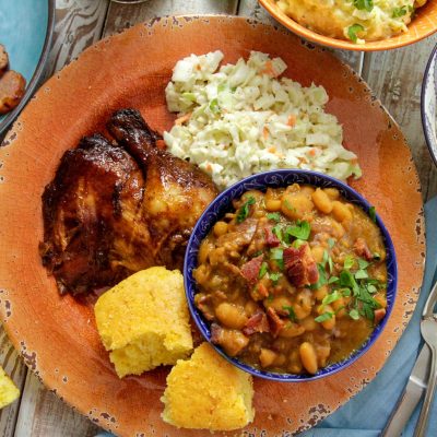 a plate of baked beans and bbq chicken with a side of coleslaw and corn bread