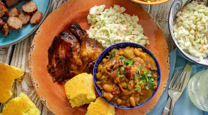 a plate of baked beans and bbq chicken with a side of coleslaw and corn bread