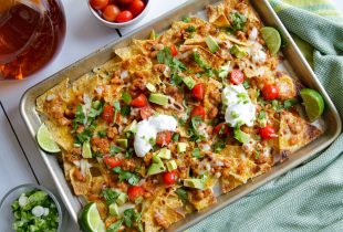 a tray of nachos topped with beans, queso, cilantro, cherry tomates, avacado with 2 dollops of sour creme and cut limes on the side