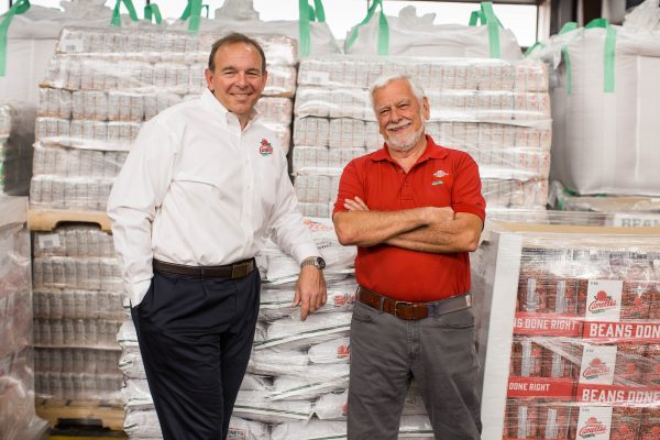 2 founders of camellia beans in front of pallets of beans