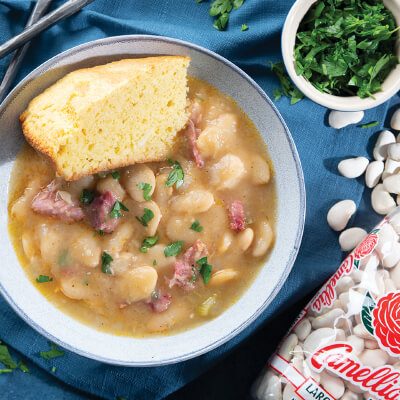 a bowl of Southern Large Limas and sausage with a side of corn bread on top next to a package of camellia brand large lima beans