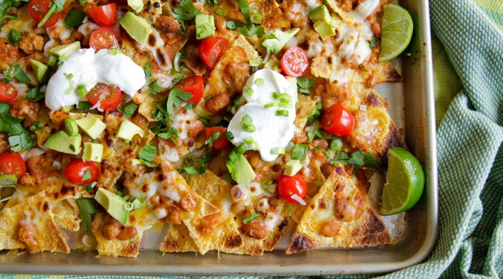 a pan of baked naked with beans, chilli, avacado, tomatoes, sour cream, and cut limes