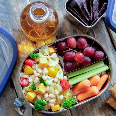 white beans and pasta with corn, broccoli and tomatoes ina lunch box with grapes and celery and carrots as a side