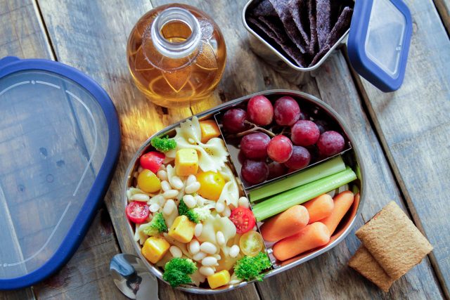 white beans and pasta with corn, broccoli and tomatoes ina lunch box with grapes and celery and carrots as a side