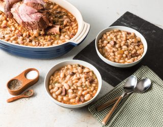 a close up of a roaster pot with white beans and a hamhock and 2 bowls full of the beans