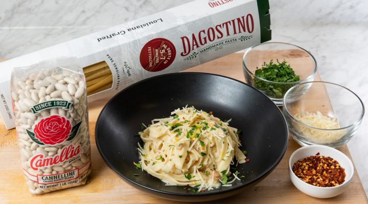 black bowl with cooked Creamy White Bean and Leek Fettuccine Alfredo surrounded by bag of beans and box of pasta and small bowls of other ingredients