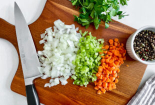 A wooden chopping block with a knife resting on the side with a variety of chopped vegetables