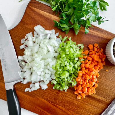 A wooden chopping block with a knife resting on the side with a variety of chopped vegetables