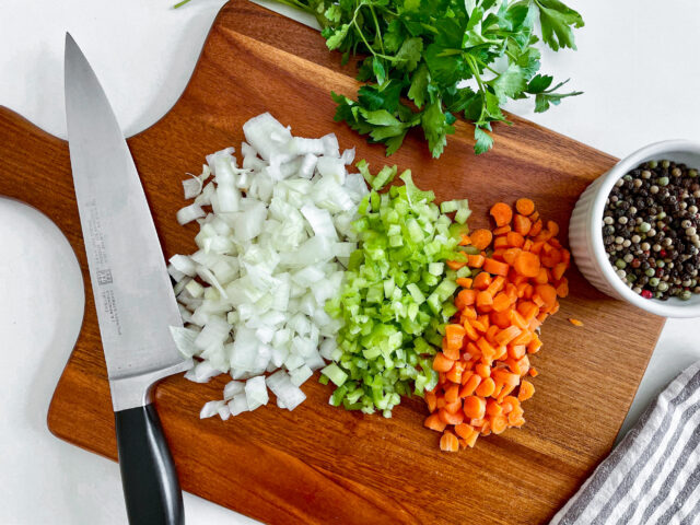 A wooden chopping block with a knife resting on the side with a variety of chopped vegetables