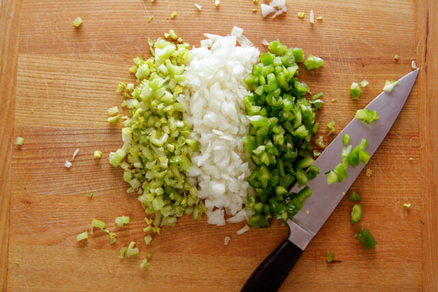 A wooden chopping block with a knife resting on the side with a variety of chopped vegetables