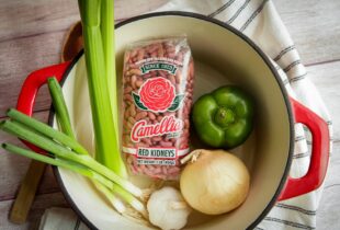 a top down view of a dutch oven with a package of red beans, slices of celery,a green bell pepper, an onion and a garlic clove