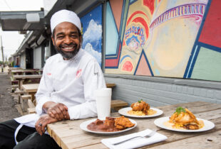 chef smiling and sitting outside heard dat resturant in from of their 3 more popular dishes