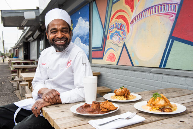 chef smiling and sitting outside heard dat resturant in from of their 3 more popular dishes