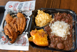 a top down view of a to go box full of red beans and rice with a side of corn bread, mac and cheese and fried chicken