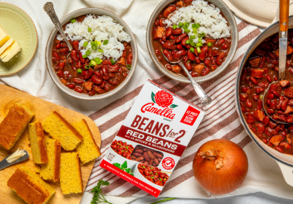 Two bowls of red beans and rice topped with green onions, beside a Camellia Beans for Two package on a striped cloth with corn bread slices nearby.