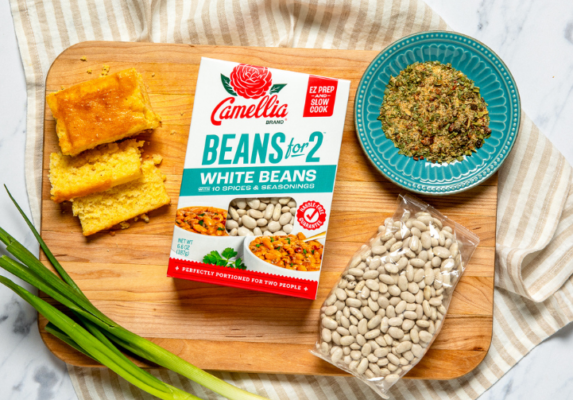 Wood cutting board with cornbread squares, a box of Camellia Beans for Two White Beans, a bag of white beans, a teal plate of crumbed topping, and green onions.