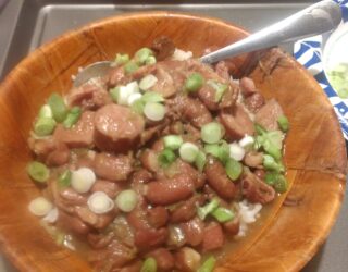 A wooden bowl filled with red beans, sliced sausage, and chopped green onions served over white rice, with a spoon in the bowl. In the background are tomatoes, an apple, and other food items.