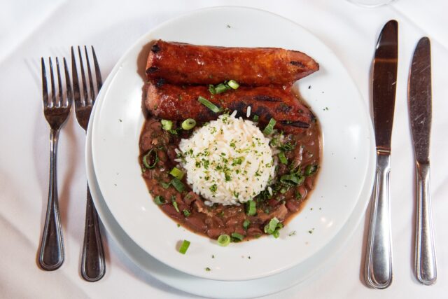 Overhead shot of white bowl full of Galliano Restaurant’s Red Beans and Rice and 2 large pieces of andouille sausage, on white, cloth tablecloth