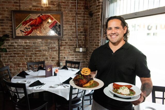 Galliano’s Chef holds a plate of their Pork Chop LaPlace entree and a bowl of their Red Beans and Rice entrees inside the restaurant’s dining room, with exposed brick wall behind him