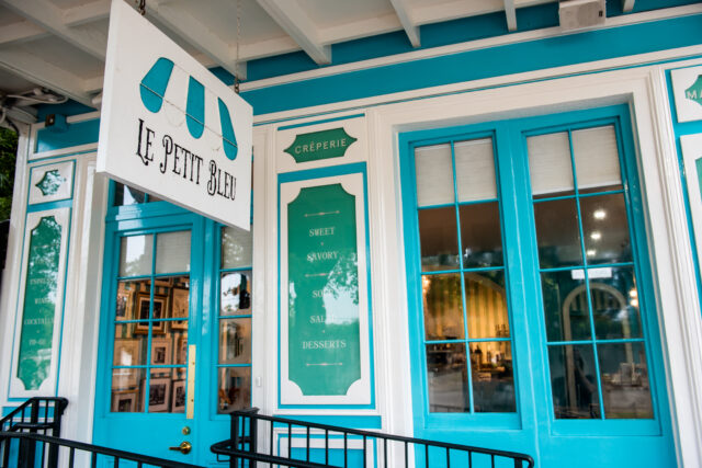 Exterior of a vibrant café with teal and white accents. A sign reading "Le Petit Bleu" hangs above a blue-framed window and door, creating a cozy, inviting atmosphere.