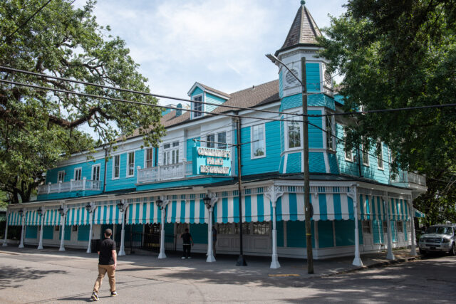 A vibrant turquoise Victorian-style building with white trim and striped awnings, surrounded by trees. Two people walk nearby under a sunny sky.