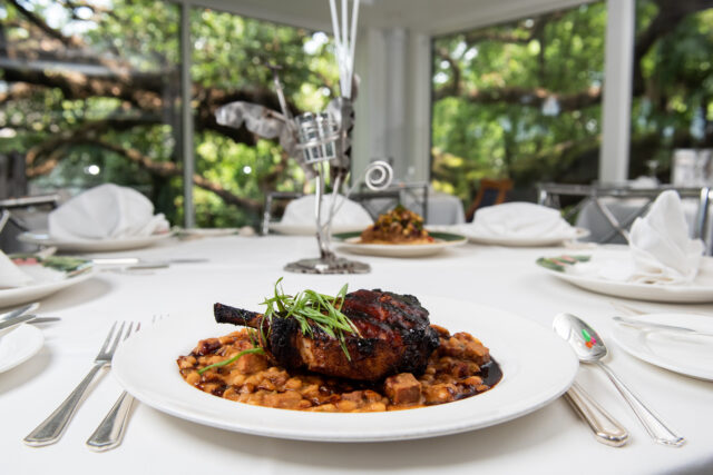 Grilled meat with sauce and beans on a set table, surrounded by elegant cutlery and napkins. Large windows reveal a lush, green garden backdrop.