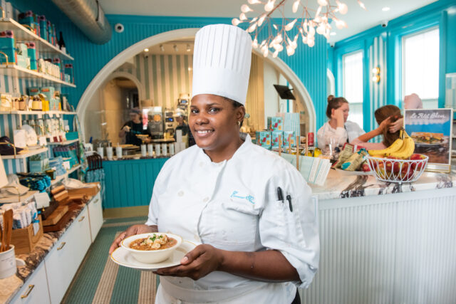 Smiling chef in a white uniform and tall hat holds a bowl of soup in a bright, turquoise-themed bakery. Ingredients and décor create a cheerful, inviting atmosphere.