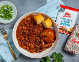 A bowl of hearty with beans and a glazed chicken drumstick, served with cornbread. Nearby are green onions, a fork, and a bean flavor infusion packet.