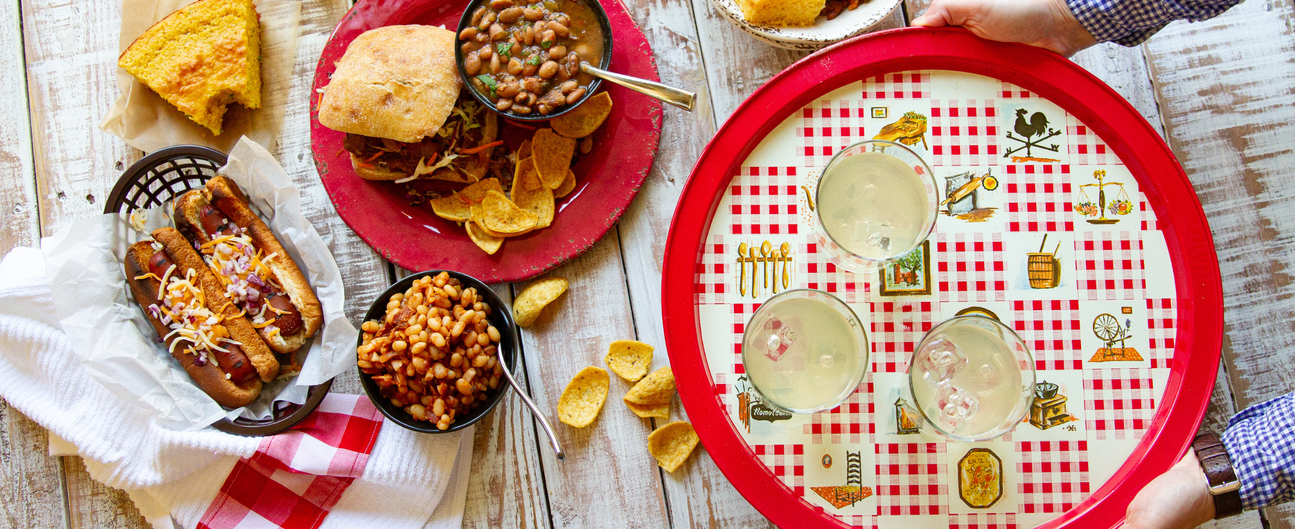 A rustic table with a hot dog basket topped with cheese, a sandwich with baked beans and corn chips, cornbread, and a red tray holding three drinks.