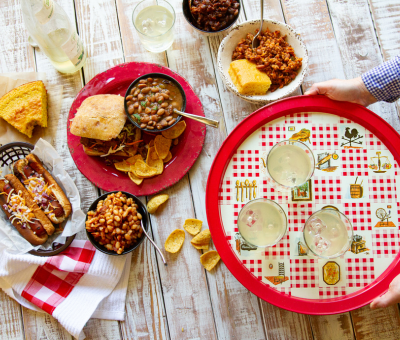 A wooden table with hot dogs topped with cheese, a sandwich with baked beans and corn chips, cornbread, and a red tray holding three drinks.