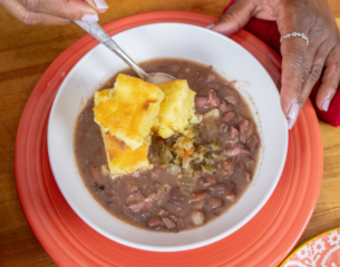 A person holds a bowl of hearty red bean soup with a spoon and cornbread, on a red plate. The scene conveys warmth and comfort.