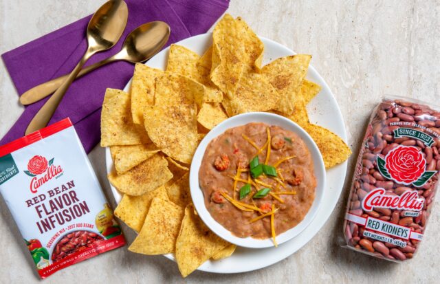 A white plate of tortilla chips surrounds a bowl of bean dip topped with cheese and green onions. Nearby, red bean infusion seasoning and a bag of red kidney beans are displayed. A purple napkin and gold spoons add elegance.