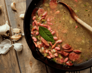A rustic wooden table holds a cast iron pot filled with sausage and bean soup. Garlic cloves are scattered beside it, and a wooden spoon is in the pot.