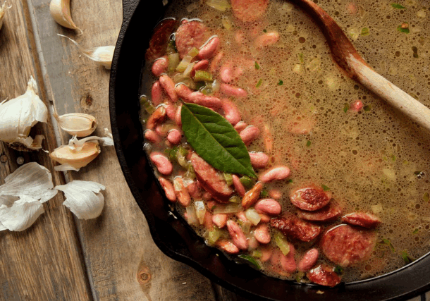 A rustic wooden table holds a cast iron pot filled with sausage and bean soup. Garlic cloves are scattered beside it, and a wooden spoon is in the pot.
