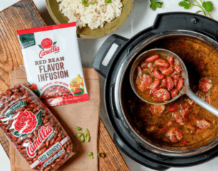 Overhead view of a pot with red beans and sausage stew. Nearby are packages of Camellia red kidney beans and flavor infusion. A bowl of rice sits beside.