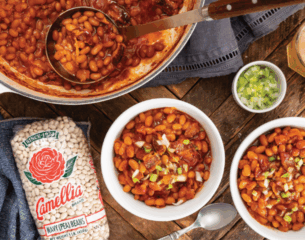 Bowl of baked beans with bacon and green onions, surrounded by a bag of navy beans and a pot. Rustic setting with a cozy, home-cooked feel.