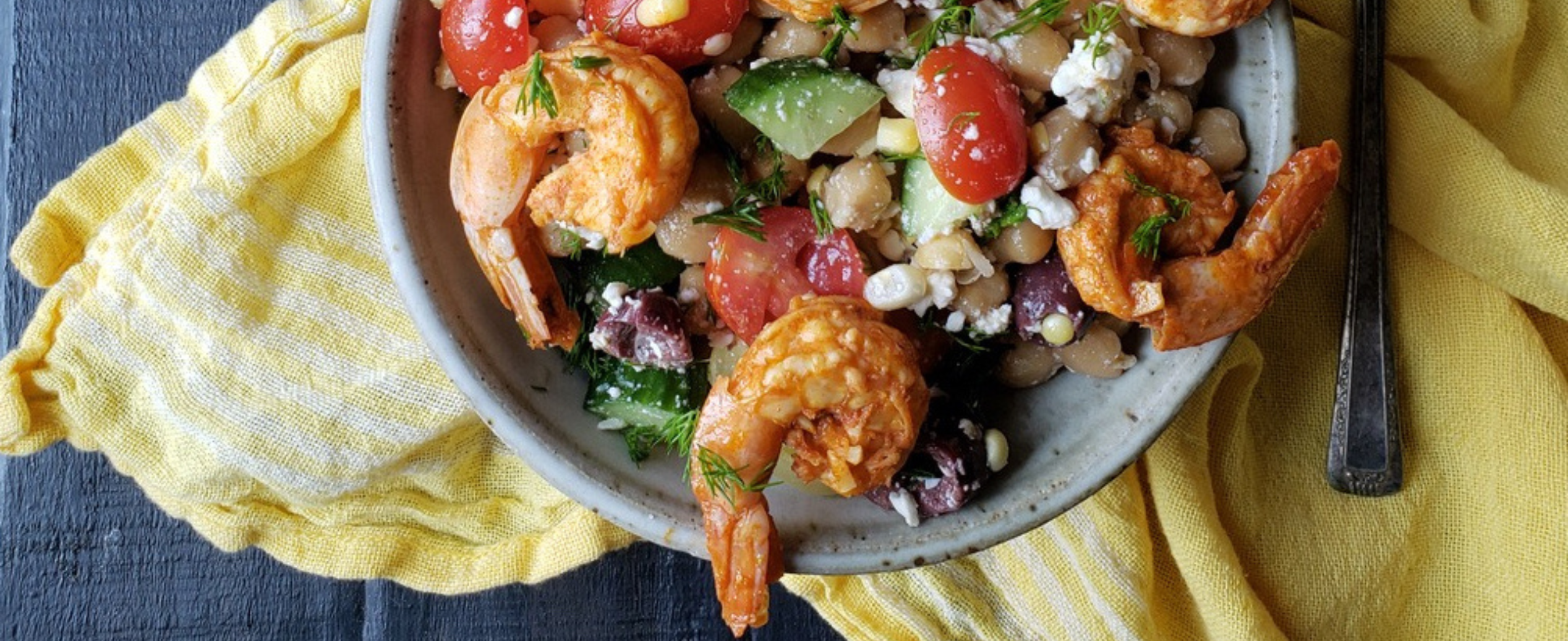 A bowl of shrimp salad with cherry tomatoes, cucumbers, olives, and feta on a yellow cloth. The setting is rustic with a dark wood table.