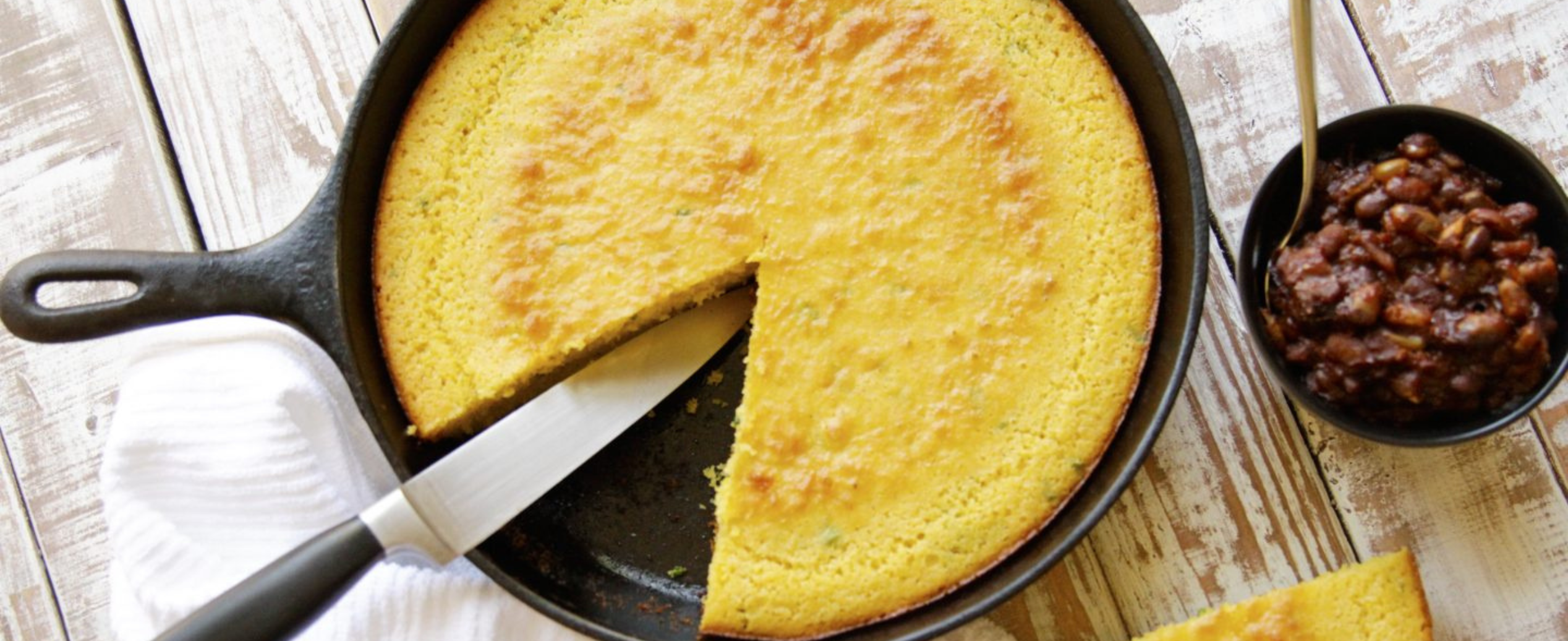 A golden cornbread in a cast iron skillet has a wedge cut out. A knife rests on the edge. A bowl of baked beans is beside the skillet on a rustic table.