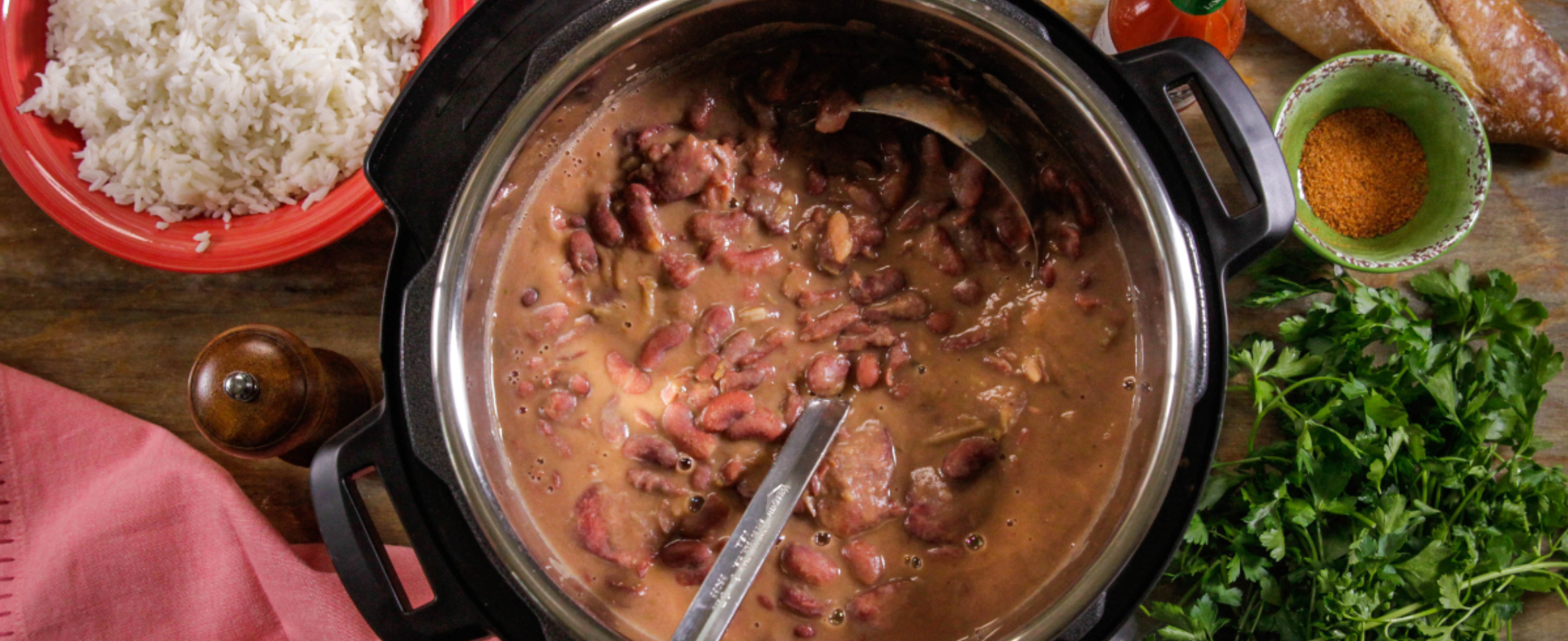 A pot of red beans with a ladle sits on a wooden table, surrounded by cooked rice, a baguette, spice bowl, fresh parsley, hot sauce, and a pepper mill.