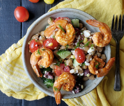 A bowl of shrimp salad with cherry tomatoes, cucumbers, olives, and feta on a yellow cloth. The setting is rustic with a dark wood table.