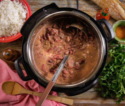 A pot of red beans with a ladle sits on a wooden table, surrounded by cooked rice, a baguette, spice bowl, fresh parsley, hot sauce, and a pepper mill.