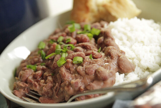 A white plate with red beans garnished with green onions next to white rice, conveying a warm, hearty meal. A bread roll is in the background.